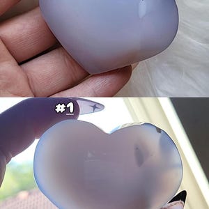 May include: Two heart-shaped stones in shades of blue and white. The top stone is held by a hand with a light-colored background. The bottom stone is held by a hand with the number "#1" visible. The stones have a smooth, polished surface.
