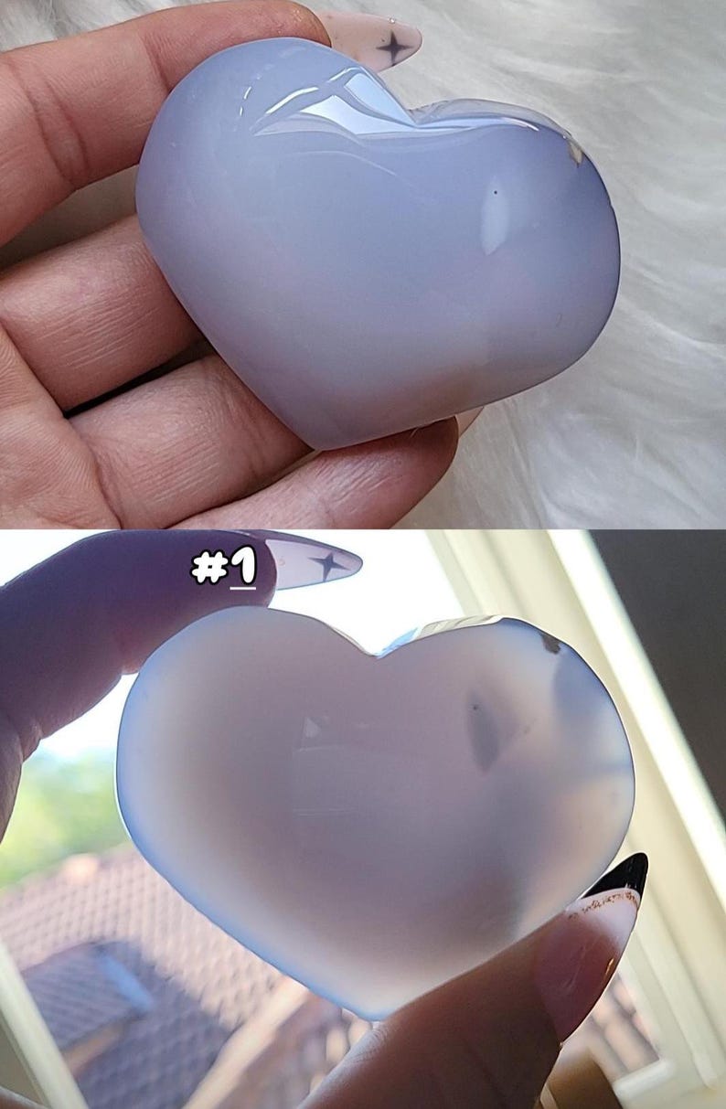 May include: Two heart-shaped stones in shades of blue and white. The top stone is held by a hand with a light-colored background. The bottom stone is held by a hand with the number "#1" visible. The stones have a smooth, polished surface.