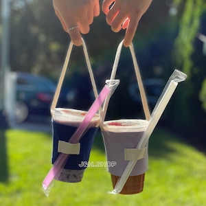 May include: Two clear plastic cups with lids and straws, each with a fabric sleeve and a white strap handle. The cups are filled with a pink and brown beverage. The text "J&L SHOP" is visible on the sleeves.
