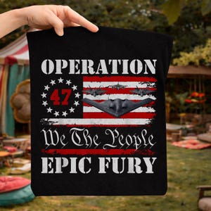 Puede incluir: Bolso tote negro con un diseño de bandera estadounidense desgastada. El texto dice "OPERATION 47", "We The People" y "EPIC FURY". La bandera incluye un bombardero B-2 y otras aeronaves.