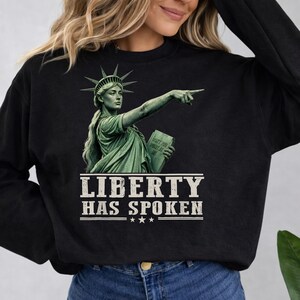 Könnte beinhalten: Ein schwarzes Langarm-Sweatshirt mit einer Grafik der Freiheitsstatue, die mit einer Hand zeigt und eine Tafel mit der Aufschrift "DON'T YOU POINT AT ME" hält. Unter der Grafik steht der Text "LIBERTY HAS SPOKEN" in Weiß.