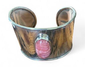 May include: A wide silver cuff bracelet with a polished, wood-grain-like texture. The bracelet features a large, oval, pink gemstone set in the center. The edges of the bracelet are rounded and smooth, creating a modern, elegant look.