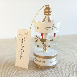 May include: White wooden carousel ornament with a gold painted base and a white horse. The ornament has a gold string attached to it and a tag that says "Thank You".