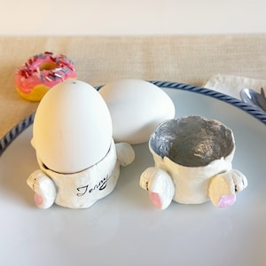 May include: Two white ceramic bunny-shaped egg holders with pink accents, one holding a white egg. The holder has the name "Jenny" written on it. A white egg and a pink donut are also on the plate.