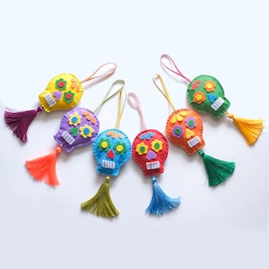 以下が含まれることがあります： カラフルなフェルト製のスカルオーナメントのコレクション。それぞれユニークなデザインです。スカルには、花や縫い付けられた口などの刺繍が施され、下にはタッセルがぶら下がっています。オーナメントは、黄色、紫、青、赤、オレンジ、緑など、さまざまな色があります。