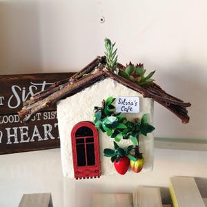 May include: A miniature house with a cream-colored facade, a red arched door, and a twig roof. The house is decorated with green leaves, red and yellow fruit, and a sign that reads "Silvia's Cafe". A wooden sign with text is visible in the background.