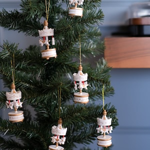 May include: A Christmas tree decorated with white and gold carousel horse ornaments. The ornaments feature a red roof and gold trim.