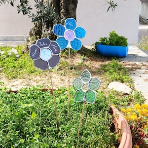 以下が含まれることがあります： ステンドグラスで作られた、カラフルな花のガーデンピックが3つ。1つは青、1つは緑、もう1つは青と茶色の混合色です。それぞれの花には名前が書かれています。ガーデンピックは庭のベッドに立っています。