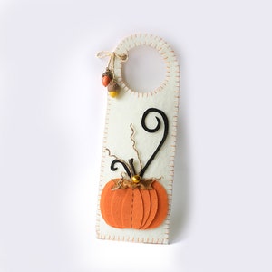 May include: Cream-colored felt door hanger with an orange pumpkin applique and black stem detail. The hanger is stitched with orange thread and adorned with hanging acorns. A festive autumn decoration.