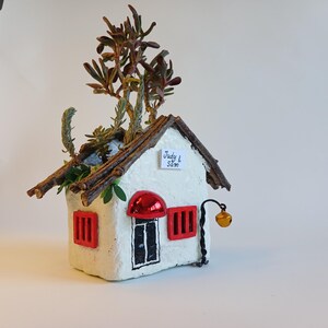 May include: A handcrafted white house-shaped planter with a textured finish, featuring a brown twig roof and red accents on the windows and door. The planter is filled with various green and brown succulents. A small sign reads "Judy & Sam".