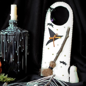 May include: A white door hanger with a black witch's hat, a broom, and bats. The hanger is decorated with gold glitter and a purple bead. A black bottle with white wax dripping down the side is in the foreground.