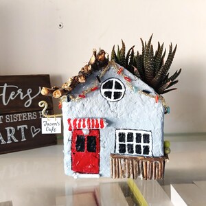 May include: A handmade, light blue miniature house planter with a red door and awning, a round window, and a rectangular window. The roof is decorated with twigs and a succulent plant. A small sign reads "Jason's Cafe."