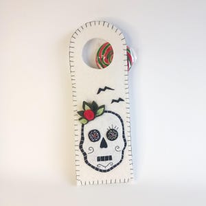 May include: White felt door hanger with a black embroidered skull design. The skull has a red flower and black eyes. The hanger has a cut-out circle at the top.