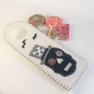 May include: White felt door hanger with a black and white sugar skull design, a grey top hat, and a black stitched border. The door hanger has a round opening at the top. Three lollipops are visible in the background, one green and white, one red, and one watermelon.