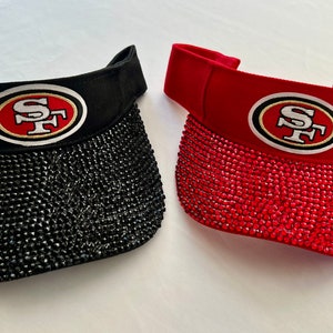 May include: Two visors, one black and one red, each adorned with sparkling embellishments. The visors feature the San Francisco 49ers logo in the center, with a red and gold emblem on a white background.