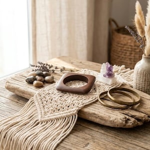 May include: A cream-colored macrame wall hanging with a diamond pattern and fringe, featuring a square dark brown wooden bangle, two thin brass-toned metal bangles, a cluster of smooth grey stones, and a small purple crystal point.