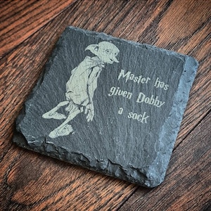 May include: A black slate coaster featuring a white image of a house elf and the text "Master has given Dobby a sock."