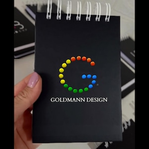 Peut inclure: Un carnet à spirales noir arborant un logo circulaire composé de petits points multicolores disposés en forme de lettre G. Sous le logo, le texte GOLDMANN DESIGN est imprimé dans une police blanche sans empattement épurée.