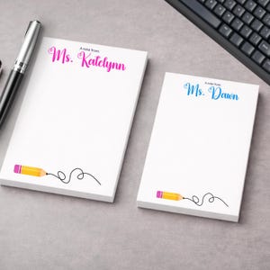 以下が含まれることがあります： パーソナライズされたテキストが書かれた2つの白いメモ帳。1つはピンク色の「Ms. Katelynn」、もう1つは青色の「Ms. Dawn」と書かれています。各メモ帳には、ピンクの消しゴムと黒い線が付いた黄色の鉛筆のグラフィックが描かれています。ペンとキーボードも表示されています。