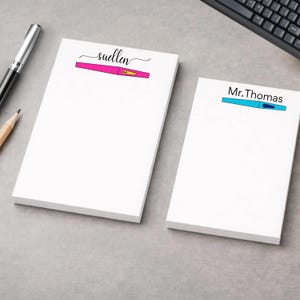 Può includere: Due blocchi per appunti bianchi con testo personalizzato. Un blocco per appunti ha il nome "suellen" sopra un'illustrazione di penna rosa e blu. L'altro blocco per appunti ha "Mr.Thomas" sopra un'illustrazione di penna blu e gialla. Sono visibili una penna e una matita.