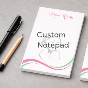 Könnte beinhalten: Ein weißer rechteckiger Notizblock mit dem Text Maison Belle in rosa Schreibschrift, den Worten Custom Notepad in Dunkelgrau, einer hellgrauen Strichzeichnung einer Person und rosa sowie grünen Wellenlinien am unteren Rand, neben einem schwarzen Stift und Bleistift.