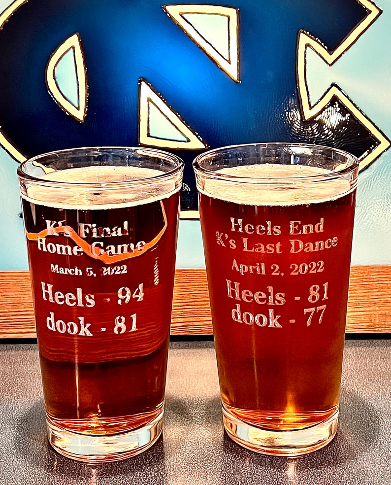Peut inclure: Deux chopes &agrave; bi&egrave;re en verre transparent remplies de bi&egrave;re brune. Les chopes sont grav&eacute;es avec du texte qui dit "Final Home Game March 5, 2022 Heels - 94 dook - 81" et "Heels End K's Last Dance April 2, 2022 Heels - 81 dook - 77".