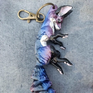 May include: A handmade fox keychain with a galaxy-themed design. The fox is painted in shades of blue, purple, and white, with black paws and a white tail tip. It has a gold-colored clasp for attaching to keys or bags.
