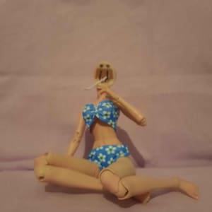 May include: A headless doll wearing a blue bikini with white floral patterns. The doll is in a seated position with its legs bent. The background is a solid light purple colour.