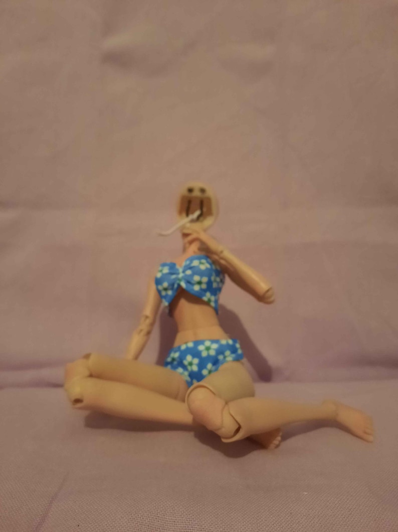 May include: A headless doll wearing a blue bikini with white floral patterns. The doll is in a seated position with its legs bent. The background is a solid light purple colour.