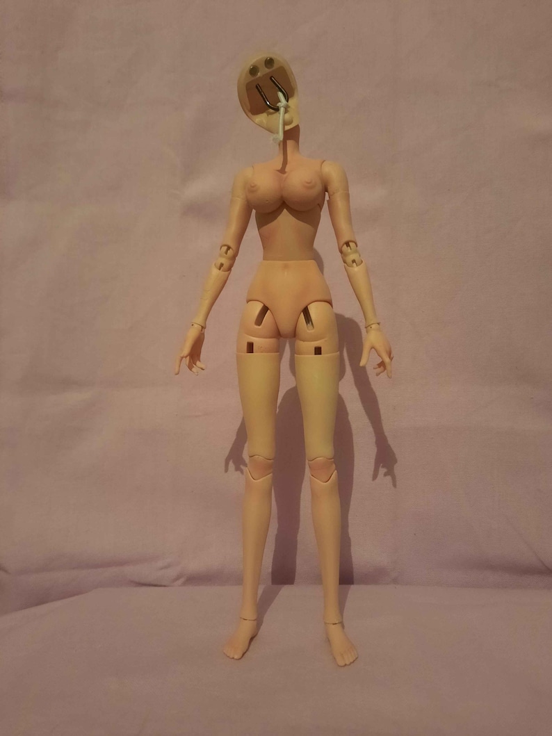 May include: A nude-coloured articulated doll with a detached head and exposed internal mechanisms. The doll has a slender build, with visible joints at the shoulders, elbows, hips, and knees. The doll's head is open, revealing internal components.
