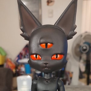 Puede incluir: Una figurita negra con forma de gato con tres ojos naranjas y un cuerpo negro. La figurita tiene una pequeña nariz puntiaguda y una pequeña boca.