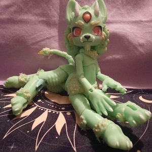 May include: A light green fantasy doll with a wolf-like head, three eyes, and small horns. The doll has articulated limbs and a long tail. The doll is sitting on a dark blue surface with a white celestial design.