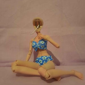 May include: A doll wearing a blue bikini with white floral patterns. The doll is missing its head and one arm. The bikini top has a bow in the centre. The doll is sitting with its legs crossed.