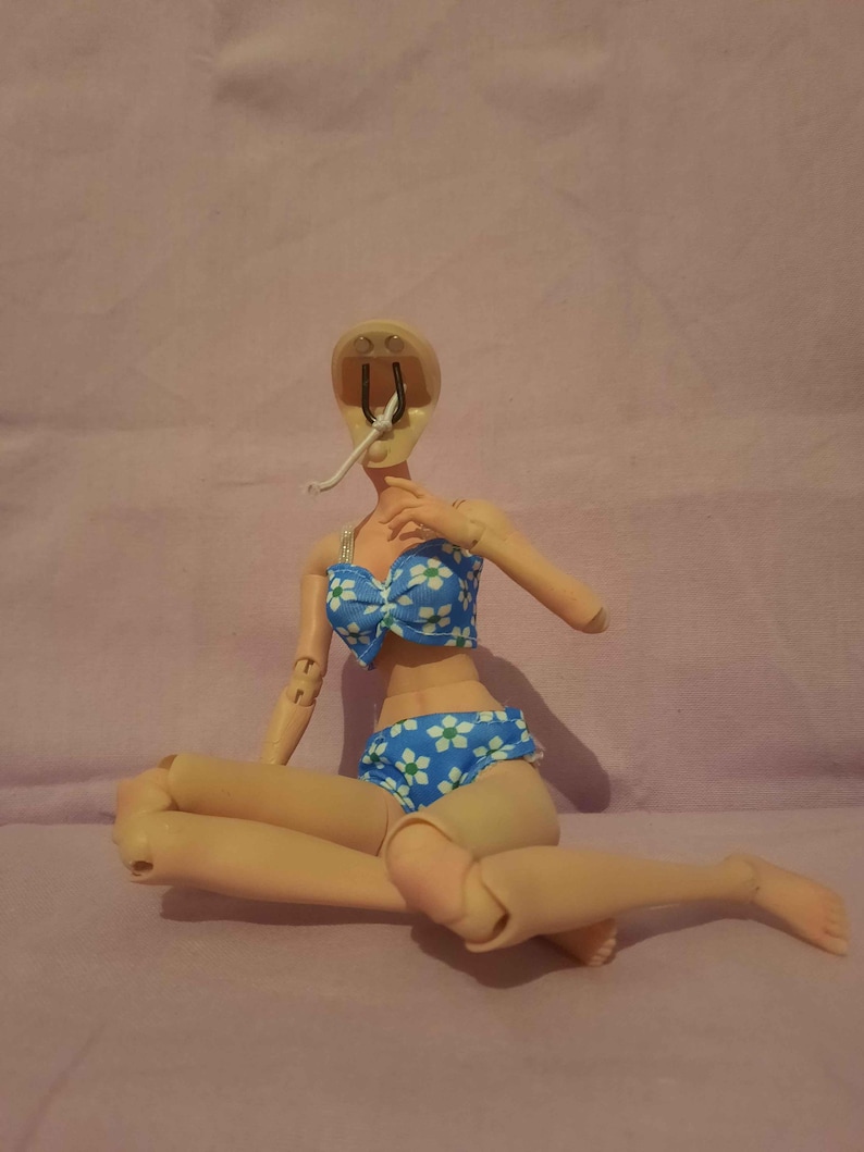 May include: A doll wearing a blue bikini with white floral patterns. The doll is missing its head and one arm. The bikini top has a bow in the centre. The doll is sitting with its legs crossed.
