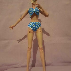 May include: A doll in a blue bikini with white floral patterns. The doll has articulated joints and a neutral skin tone. The bikini top features a bow, and the bottoms are high-waisted. The doll is posed with one arm raised.