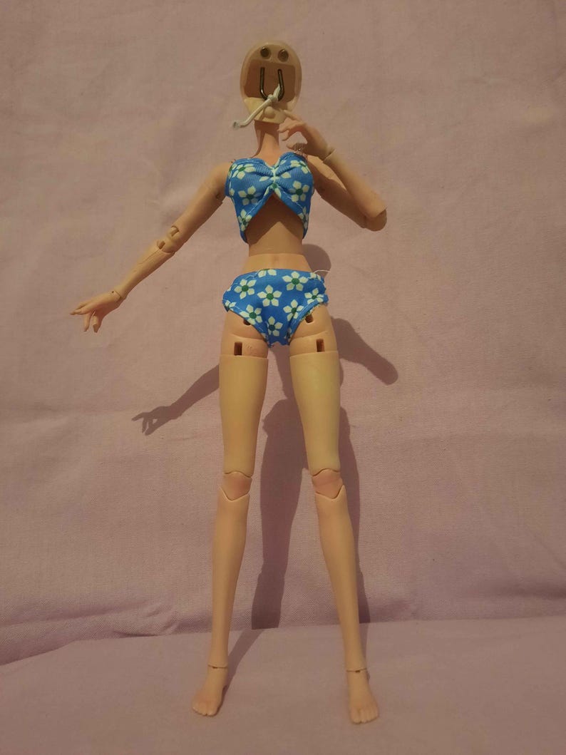May include: A doll in a blue bikini with white floral patterns. The doll has articulated joints and a neutral skin tone. The bikini top features a bow, and the bottoms are high-waisted. The doll is posed with one arm raised.