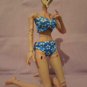 May include: A doll in a blue bikini with a white floral design. The bikini top features a bow. The doll is kneeling on a pink surface.