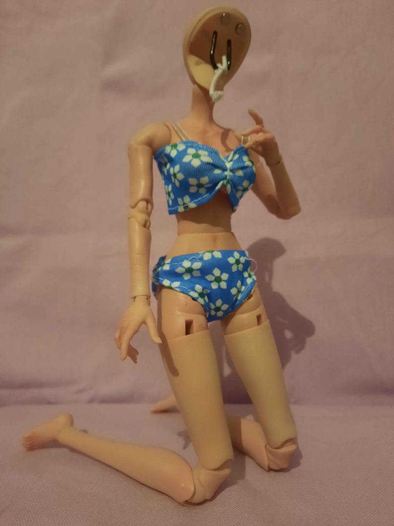 May include: A doll in a blue bikini with a white floral design. The bikini top features a bow. The doll is kneeling on a pink surface.