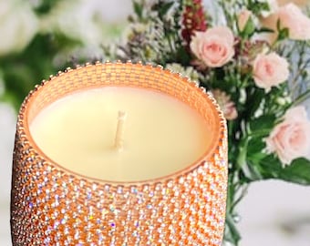 Puede incluir: Una vela decorativa en un recipiente de vidrio color cobre incrustado de joyas. La vela tiene cera de color crema y una sola mecha. El fondo presenta un arreglo borroso de rosas rosas y vegetación.