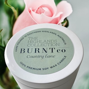 Puede incluir: Un recipiente blanco para velas con una etiqueta verde. La etiqueta dice "BURNTco Country Lane 100% Premium Soy Wax Candle". Una rosa rosa est&aacute; en el fondo. La colecci&oacute;n Highlands tambi&eacute;n est&aacute; impresa.