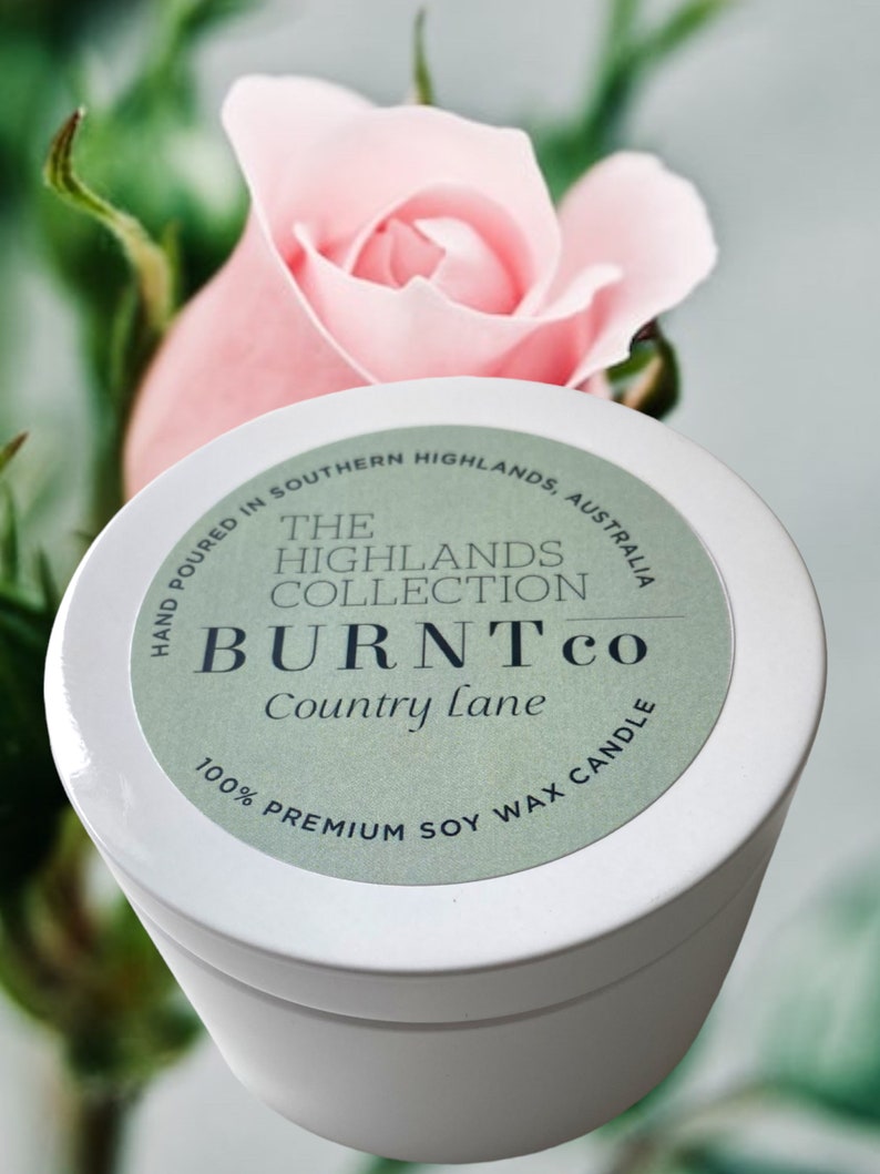 Puede incluir: Un recipiente blanco para velas con una etiqueta verde. La etiqueta dice "BURNTco Country Lane 100% Premium Soy Wax Candle". Una rosa rosa est&aacute; en el fondo. La colecci&oacute;n Highlands tambi&eacute;n est&aacute; impresa.