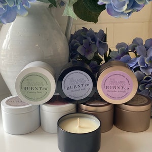 Puede incluir: Colecci&oacute;n de velas de cera de soja en latas met&aacute;licas, con etiquetas que dicen "The Highlands Collection BURNTco". Las velas est&aacute;n en latas blancas, negras y doradas, con una vela encendida en primer plano. Flores azules y moradas en el fondo.
