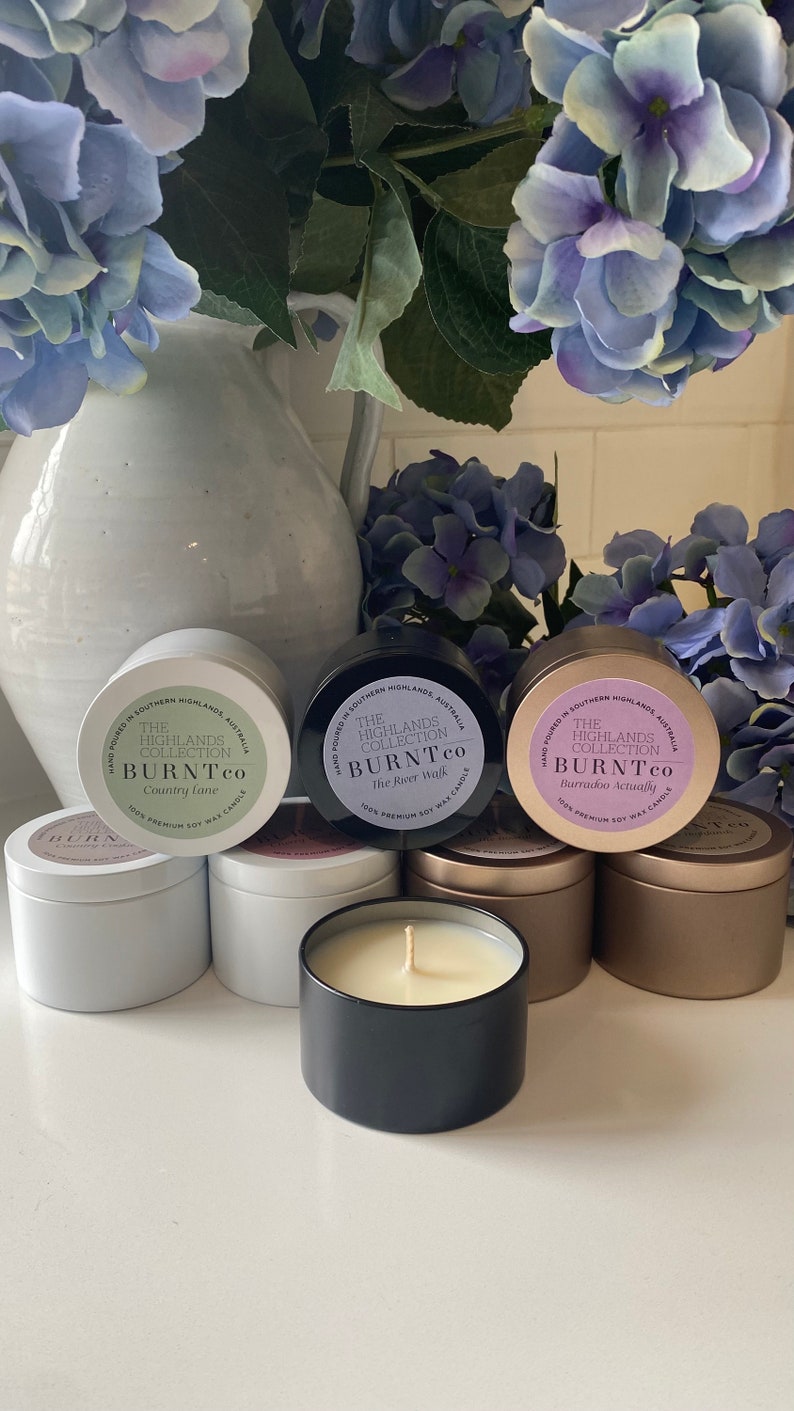 Puede incluir: Colecci&oacute;n de velas de cera de soja en latas met&aacute;licas, con etiquetas que dicen "The Highlands Collection BURNTco". Las velas est&aacute;n en latas blancas, negras y doradas, con una vela encendida en primer plano. Flores azules y moradas en el fondo.