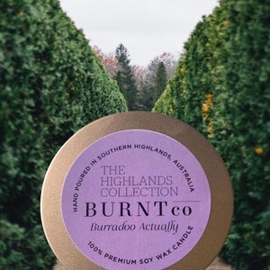 Puede incluir: Una lata de vela redonda, de color dorado, con una etiqueta morada. La etiqueta dice "The Highlands Collection BURNT co Burradoo Actually" y "100% Premium Soy Wax Candle". Tambi&eacute;n se ve el texto "Hand Poured in Southern Highlands, Australia".