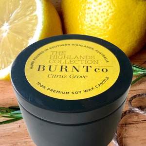 Puede incluir: Una lata de vela negra con una etiqueta amarilla que dice "BURNTco Citrus Grove 100% Premium Soy Wax Candle". La imagen incluye dos mitades de lim&oacute;n y ramitas de romero. La vela est&aacute; vertida a mano en Southern Highlands, Australia.