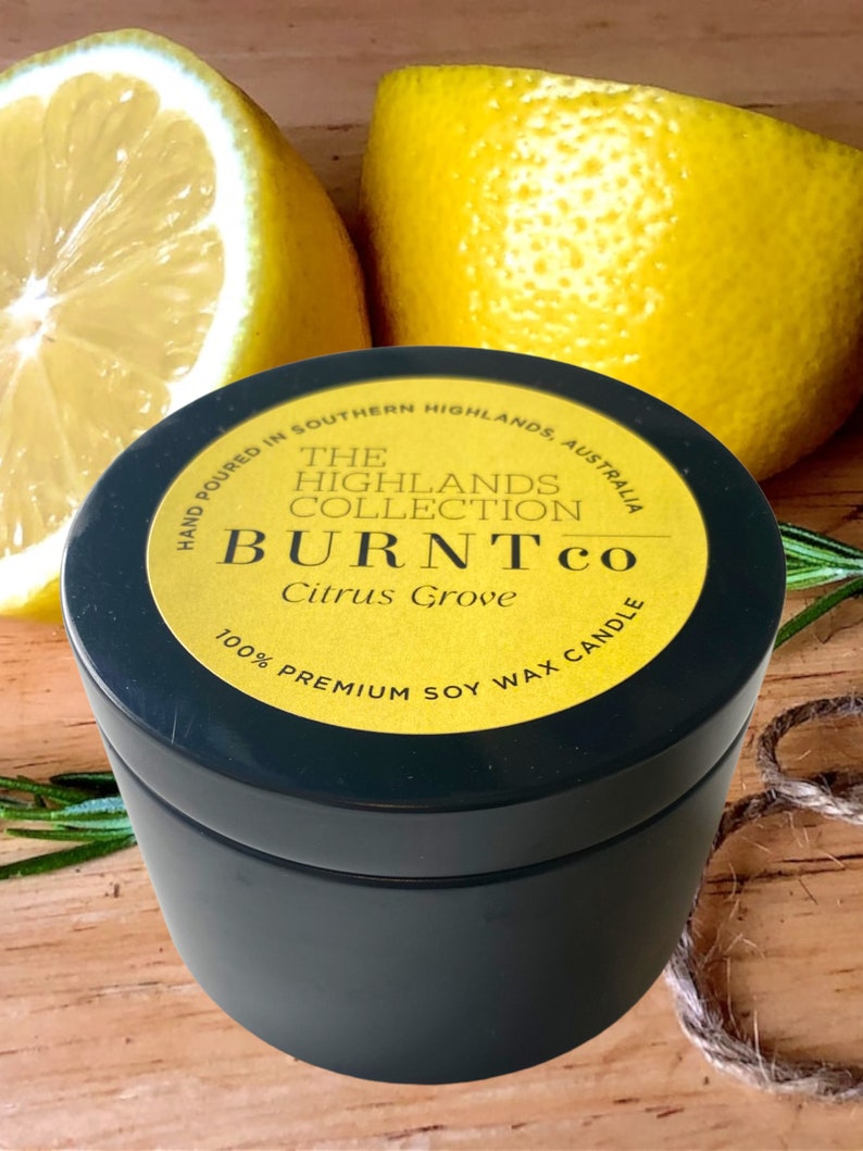 Puede incluir: Una lata de vela negra con una etiqueta amarilla que dice "BURNTco Citrus Grove 100% Premium Soy Wax Candle". La imagen incluye dos mitades de lim&oacute;n y ramitas de romero. La vela est&aacute; vertida a mano en Southern Highlands, Australia.