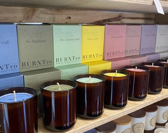 Puede incluir: Una exhibición de velas perfumadas en frascos y cajas de vidrio. Las velas están dispuestas en estantes de madera, con varios colores y aromas. Las cajas están etiquetadas como "The Highlands Collection". Los frascos son de color ámbar oscuro.