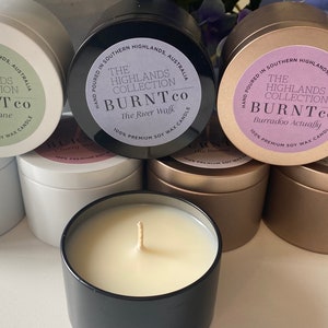 Puede incluir: Una colecci&oacute;n de velas de cera de soja en botes met&aacute;licos. Las etiquetas dicen "The Highlands Collection BURNTco" con diferentes nombres de fragancias. Las velas est&aacute;n en botes blancos, negros y dorados, con una vela encendida en primer plano.