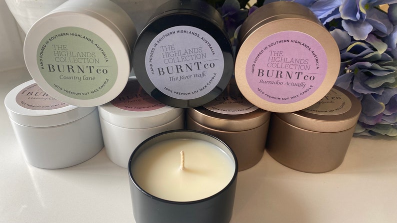 Puede incluir: Una colecci&oacute;n de velas de cera de soja en botes met&aacute;licos. Las etiquetas dicen "The Highlands Collection BURNTco" con diferentes nombres de fragancias. Las velas est&aacute;n en botes blancos, negros y dorados, con una vela encendida en primer plano.