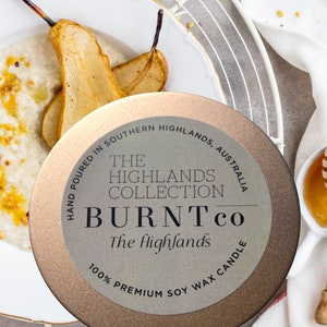 Puede incluir: Una lata de vela redonda y met&aacute;lica con el texto "BURNTco The Highlands" est&aacute; centrada en la imagen. La vela est&aacute; rodeada por un plato blanco con peras cocidas y un plato blanco con miel.